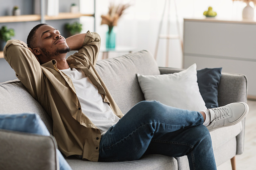 Relaxation And Comfort. Contented African American Guy Relaxing With Eyes Closed Holding Hands Behind Head Sitting On Sofa At Home. Man Enjoying Lazy Weekend In Living Room. Side View