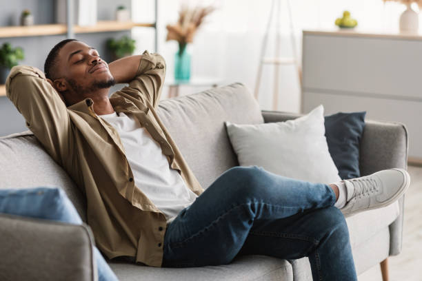 African American Guy Relaxing With Eyes Closed Sitting On Sofa Indoors Relaxation And Comfort. Contented African American Guy Relaxing With Eyes Closed Holding Hands Behind Head Sitting On Sofa At Home. Man Enjoying Lazy Weekend In Living Room. Side View taking a break stock pictures, royalty-free photos & images