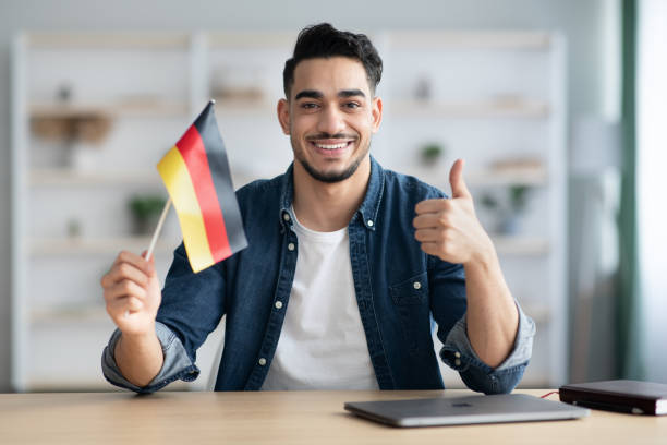 Smiling guy with flag of Germany and showing thumb up Smiling arab guy with flag of Germany showing thumb up, sitting at desk with laptop, middle-eastern young man student learning German for job, education or emmigration to Germany, copy space german language photos stock pictures, royalty-free photos & images