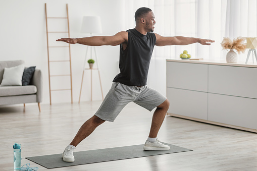 African American Man Doing Yoga Standing In Warrior Pose At Home. Black Guy Practicing Relaxing Exercises Training In Living Room Indoors. Virabhadrasana Asana. Side View Shot