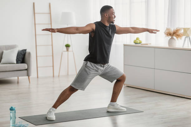 African American Man Doing Yoga Standing In Warrior Pose Indoor African American Man Doing Yoga Standing In Warrior Pose At Home. Black Guy Practicing Relaxing Exercises Training In Living Room Indoors. Virabhadrasana Asana. Side View Shot warrior position stock pictures, royalty-free photos & images
