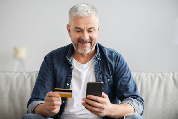 istock Smiling grey-haired man using smartphone and credit card, home interior 1322921218