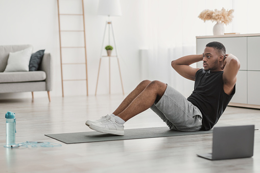 Side View Of African American Guy Doing Sit-Ups Exercising At Laptop Sitting On Floor At Home. Man Flexing Abdominal Muscles During Online Fitness Training In Living Room. Workout Concept