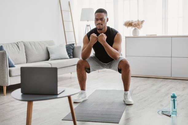 istock Sporty African Man Doing Deep Squats Near Laptop Computer Indoor 1322921358