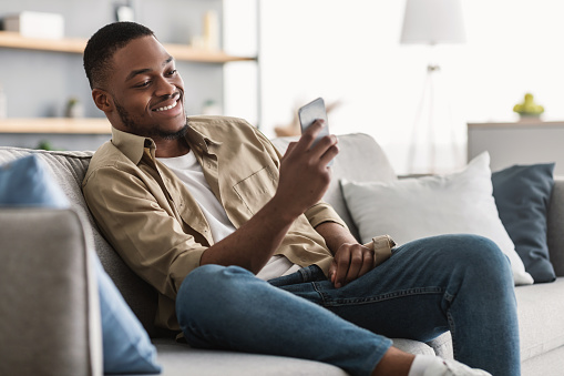 Side View Of African American Guy Using Smartphone Browsing Internet Sitting On Couch At Home. Black Man Texting On Cellphone Or Using New Application On Mobile Phone Indoors
