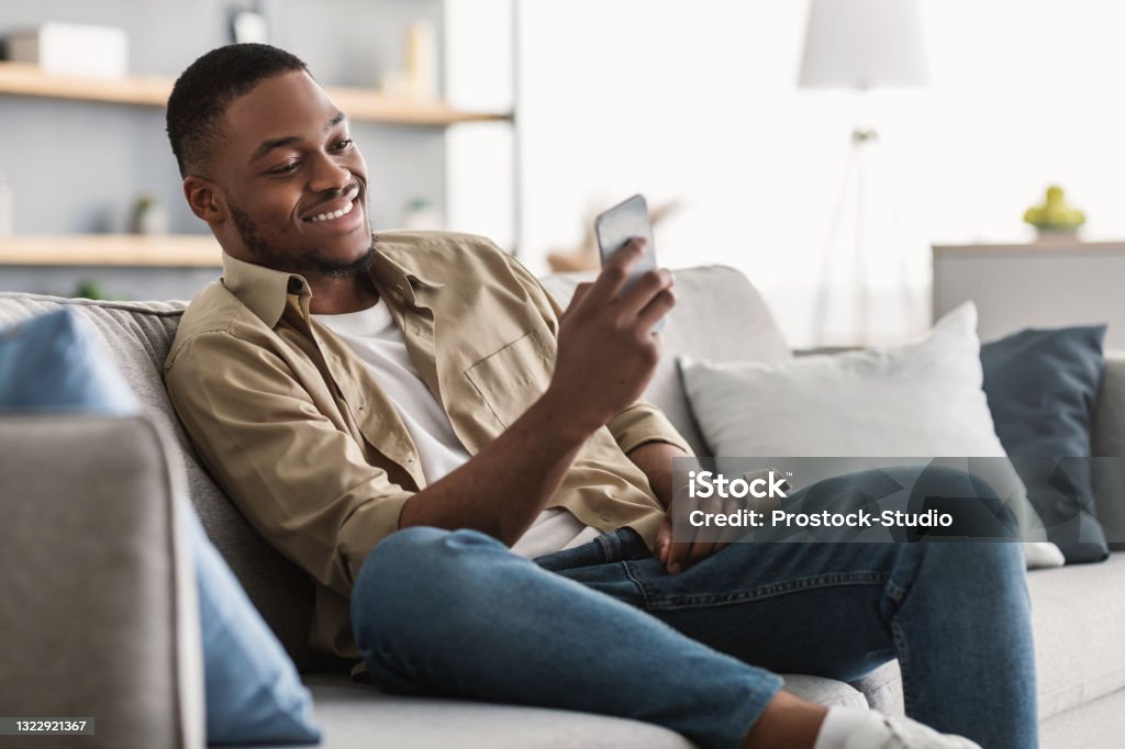 Side View Of African American Guy Using Smartphone At Home Side View Of African American Guy Using Smartphone Browsing Internet Sitting On Couch At Home. Black Man Texting On Cellphone Or Using New Application On Mobile Phone Indoors Men Stock Photo