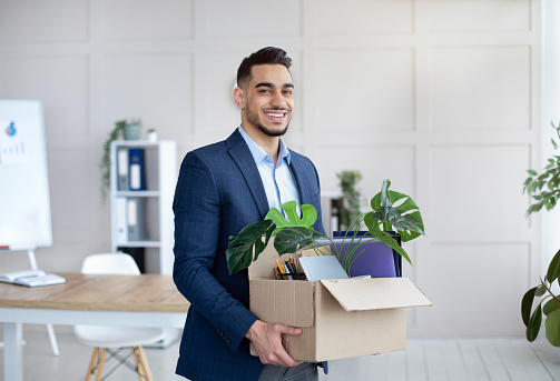 First day at office. Portrait of confident Arab businessman with belongings moving to new office. Happy young male manager holding box with supplies, changing working place, indoors