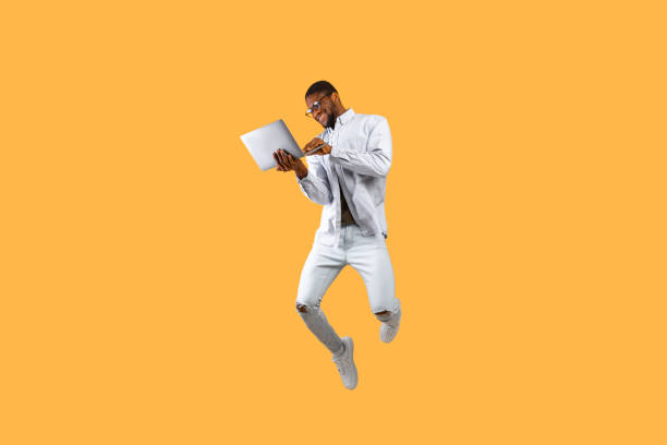 Happy african american man jumping up with laptop computer, having fun on yellow studio background