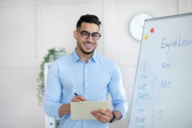 istock Positive male English teacher explaining foreign language rules near blackboard, taking notes in clipboard at office 1322922386