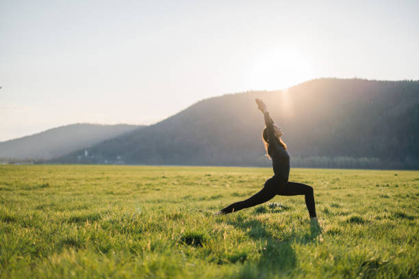 Young woman practices yoga in grassy meadow at sunrise She brings one leg forward into warrior one position, her hands over her head warrior position stock pictures, royalty-free photos & images