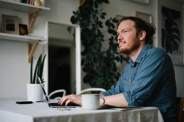 Young man works online at household kitchen table He relaxes at household kitchen table, and works on laptop rolled up sleeves stock pictures, royalty-free photos & images