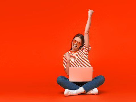 Winner! Excited smiling girl sitting on floor with laptop, raising one hand in the air is she wins, isolated on red background