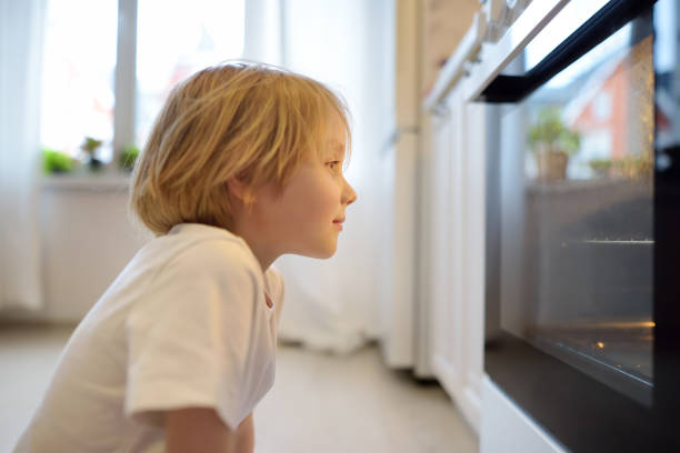 garçon heureux assis sur le sol près de la cuisinière et attendant qu’une tarte ou d’autres produits de boulangerie soient préparés. les enfants adorent la nourriture préparée par leur mère. cuisine domestique - little cakes photos et images de collection