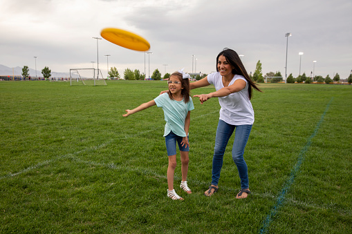 A mother shows her daughter how to throw a flying disc in a public park on a summer day.