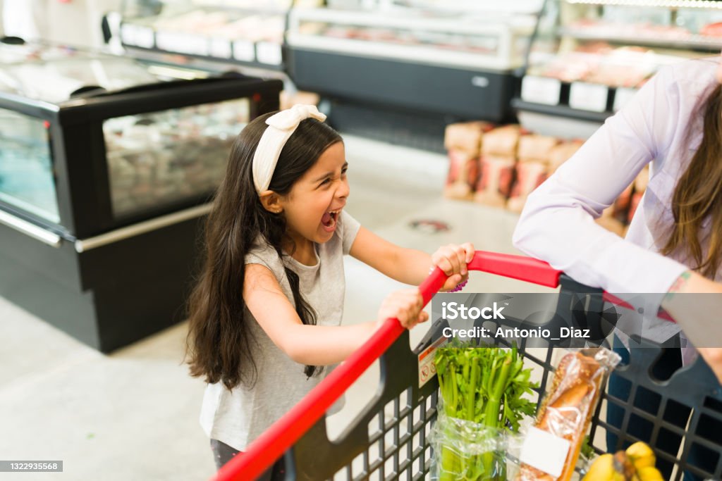 Annoyed young daughter shouting in the store Angry little kid screaming and throwing a tantrum while grocery shopping with her mom at the supermarket because she won't buy her candy Child Stock Photo