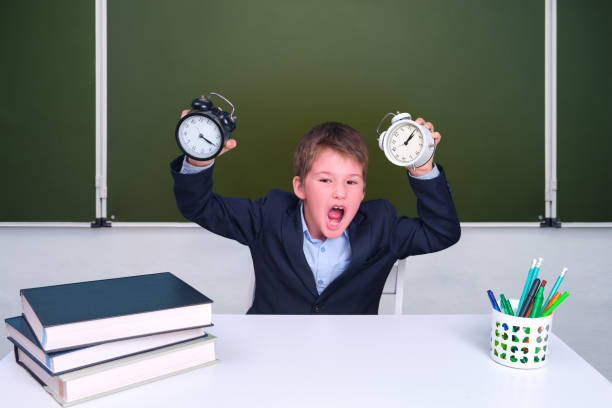 Funny junior boy with alarm clocks in the school class. Schoolboy screams at his desk at the blackboard, copy space Funny junior boy with alarm clocks in the school class. Schoolboy screams at his desk at the blackboard, copy space blackboard child shock screaming stock pictures, royalty-free photos & images