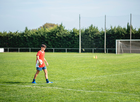 14 year old male footballer walking on sports field carrying ball before start of practice.