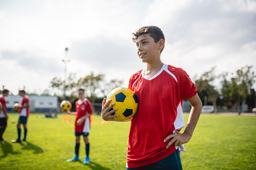 14 year old boy athlete wearing red jersey, holding soccer ball, and looking away from camera with hand on hip.
