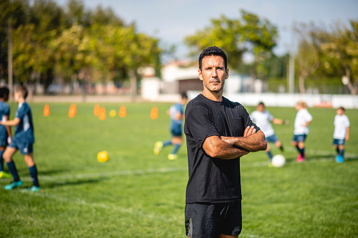 Mature male coach standing with arms crossed and looking at camera on sports field as young athletes practice in background.
