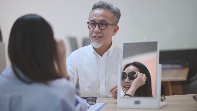 asian chinese working senior man explaining to his female customer trying on eyeglasses in front of mirror at optician store