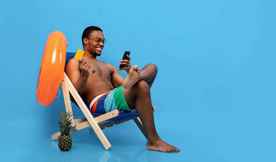 Online summer sale. Carefree black man holding credit card and smartphone, shopping on web in lounge chair, blue background, panorama with empty space. Handsome young guy purchasing goods on internet