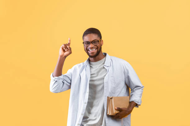 tolle idee. aufgeregt eamerikanische männliche student hält bücher und zeige mit dem finger nach oben auf gelben studio-hintergrund - male education college student nerd stock-fotos und bilder