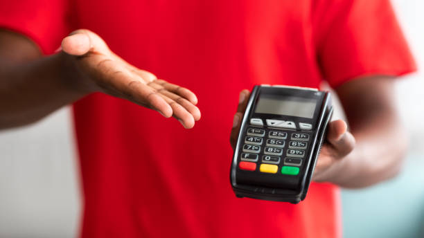 Closeup view of black man holding POS machine for payment Digital Contactless Transaction. Cropped closeup view of African American man in red t-shirt uniform holding and showing POS machine, selective focus on payment dataphone terminal, blurred background t shirt transfer stock pictures, royalty-free photos & images