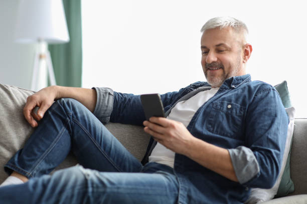 istock Smiling grey-haired mature man chilling on couch, using mobile phone 1322963817