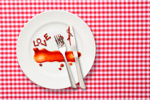 Overhead shot of word LOVE written in ketchup on a white plate after a meal, on gingham tablecloth.