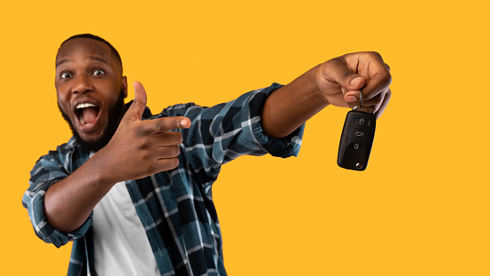 New Car. Excited African American Guy Showing His Auto Key In Excitement Posing In Studio On Yellow Background. Emotional Black Man Celebrating Buying Own Automobile. Panorama, Selective Focus