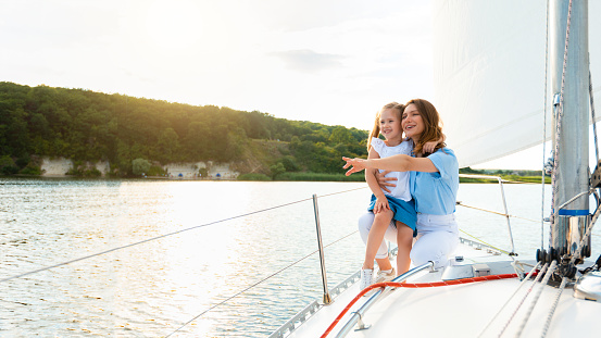Cheerful Mother And Daughter Enjoying Sailing During Yacht Ride Tour Standing On Sailboat Deck And Pointing Finger Aside. Tourism, Dream Vacation Concept. Panorama With Copy Space