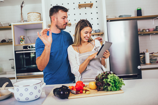 Young happy couple is enjoying and preparing healthy meal in their kitchen and reading recipes on the digital tablet.