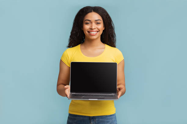 istock Positive black woman holding and showing laptop computer with black empty screen for mockup, blue background 1322973541