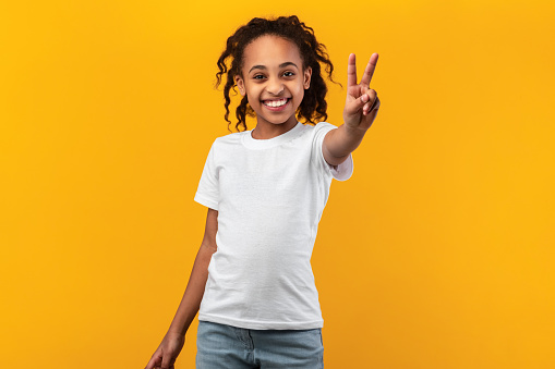 Celebrating Success. Portrait of young African American girl with curly hair wearing casual t-shirt standing and showing two fingers doing victory v sign, looking to the camera. Number 2