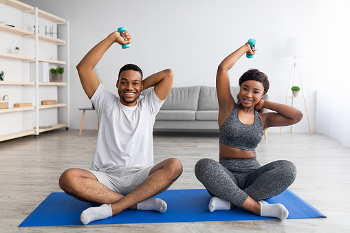 Cheerful black couple training with dumbbells at home, doing domestic fitness during covid lockdown. Strong young guy and his girlfriend training their biceps musles, keeping fit and healthy