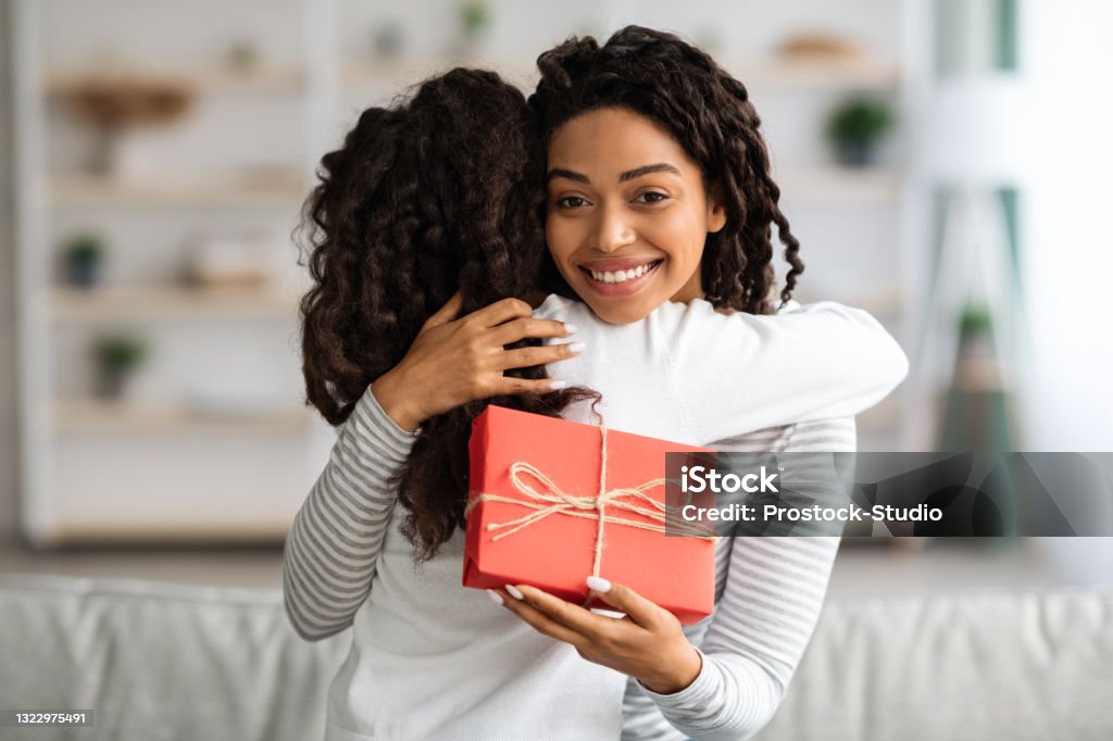 Curly black girl greeting her mom with birthday Curly black girl making birthday surprise for her excited mom, home interior, copy space. Happy african american woman holding gift box and hugging her curly daughter, smiling at camera Mother Stock Photo
