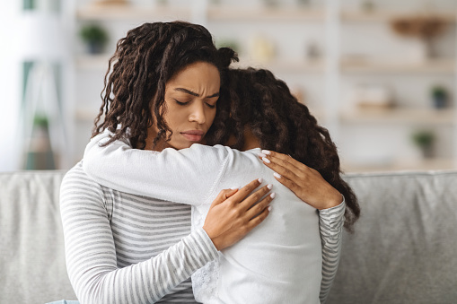 Little black girl crying on loving mothers shoulder, sitting together on sofa in living room, copy space. Closeup of african american young mom hugging her upset daughter. Motherhood concept