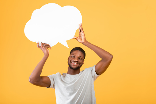 Excited african american man holding empty speech bubble above his head, posing over yellow background, studio shot. Mockup with free space for your ad or design