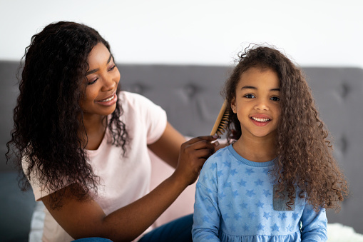 Beautiful black woman brushing her daighter's hair on bed indoors. Mother and her kid having spa day, styling their locks, spending fun family time together. Home beauty salon