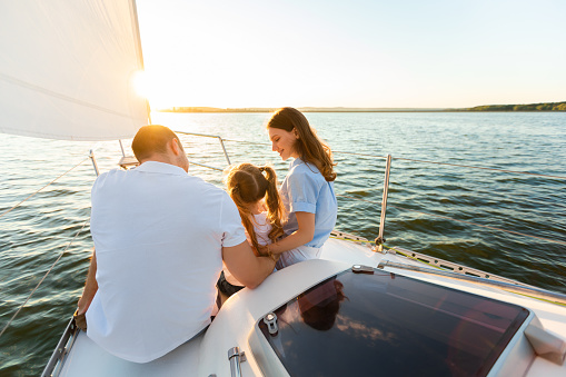 Rear View Of Family Sailing On Yacht Sitting Together On Sailboat Deck And Embracing Enjoying Sea Ride Outdoor. Unrecognizable Parents And Little Daughter Enjoying Summer Weekend On Boat