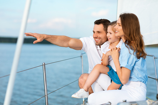Family Sailing On Yacht. Parents And Daughter Hugging Standing On Boat Deck Looking Aside At Landscapes, Enjoying Sailboat Ride Spending Summer Vacation At Seaside. Side View Shot