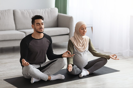 Pregnant Muslim Woman In Hijab Practicing Yoga With Her Husband At Home, Expecting Middle Eastern Spouses Meditating Together On Mat In Living Room, Sitting In Lotus Pose With Closed Eyes, Free Space