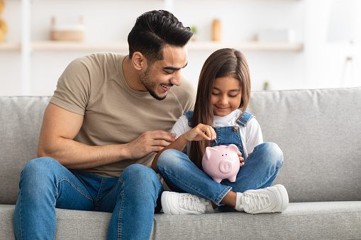 Financial Education For Children Concept. Portrait of cute smiling little girl putting coin in pink piggy bank, sitting with dad on the couch at home, man teaching his daughter how to invest