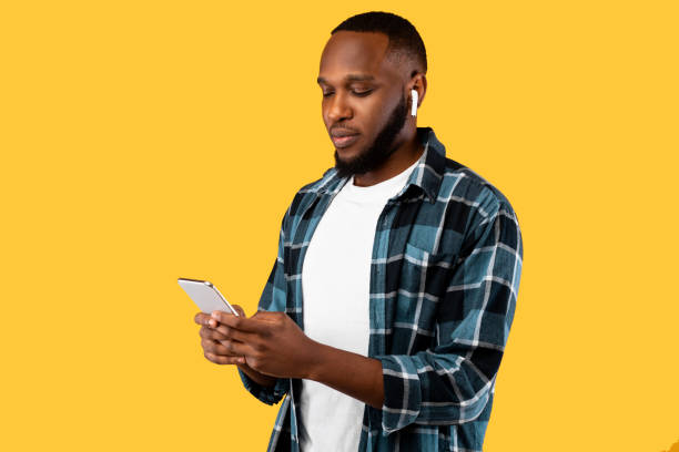istock Black Millennial Male Using Cellphone Wearing Earbuds On Yellow Background 1322978315