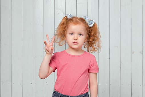 Portrait of cute ginger little girl 4-6 years old showing three fingers. Studio shot, light wooden background