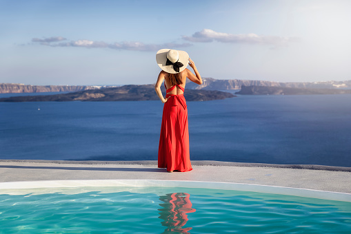 A beautiful woman in red dress and with hat stands on the edge of a swimming pool and enjoys the elevated view over the mediterranean sea during sunset time