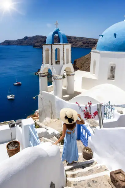 Photo of A woman in a blue dress walks down next to the blue domed church in the village of Oia, Santorini,