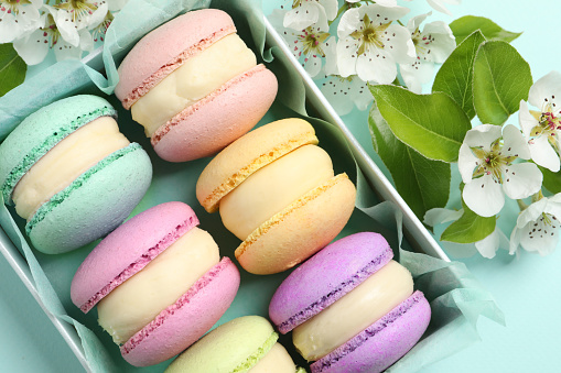 Many delicious colorful macarons in box and flowers on light blue background, flat lay