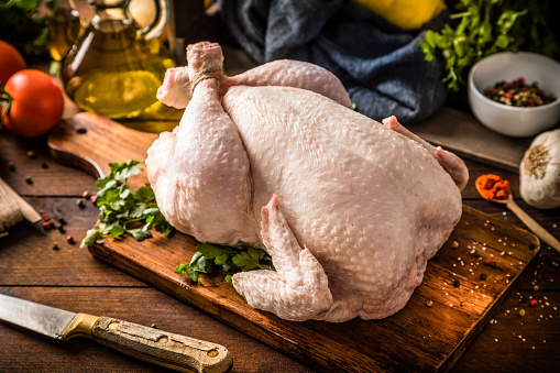 Front view of a fresh raw chicken on a rustic wooden table. The chicken is on a cutting board and is surrounded by a kitchen knife and some ingredients such as a garlic clove, an olive oil bottle, some tomatoes, and parsley.