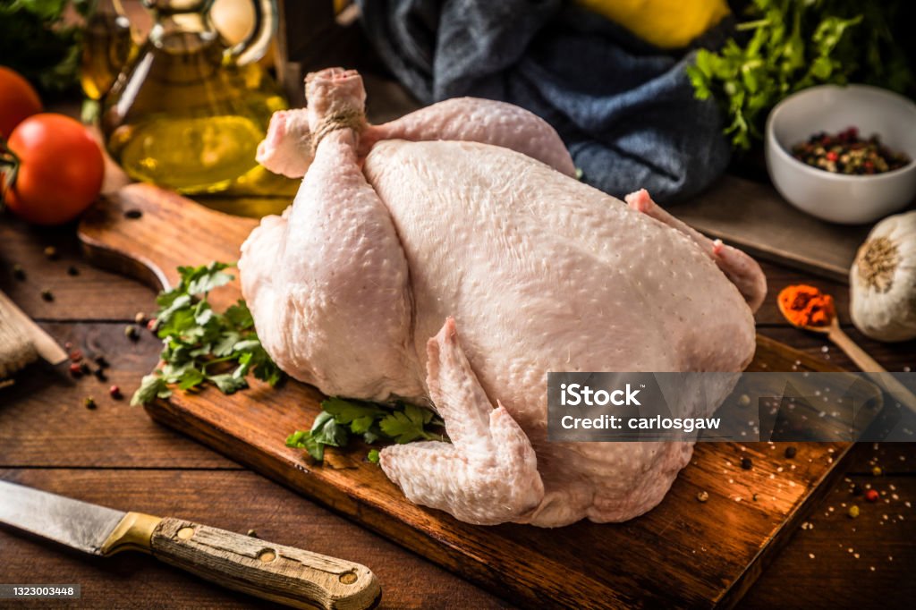 Fresh raw chicken on a rustic wooden table. Front view of a fresh raw chicken on a rustic wooden table. The chicken is on a cutting board and is surrounded by a kitchen knife and some ingredients such as a garlic clove, an olive oil bottle, some tomatoes, and parsley. Chicken Meat Stock Photo